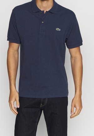 Mann trägt ein navyblaues, kurzärmliges Poloshirt mit einem kleinen grünen Krokodil-Logo auf der linken Brust und schwarze Jeans.
