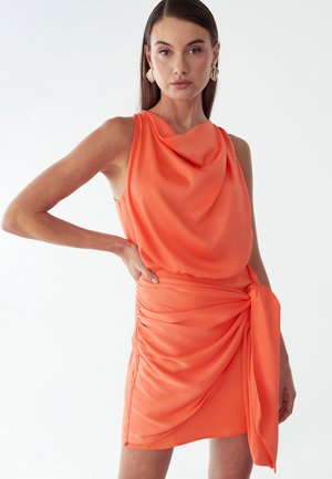 Donna che indossa un vestito corallo senza maniche con scollo drappeggiato e gonna arricciata con fiocco laterale su sfondo bianco.