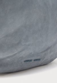 Geantă din piele intoarsă gri, cu o textură mată. Are un logo discret în relief. Formă moale, rotunjită, cu o suprafață netedă și fără elemente metalice vizibile.