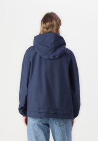 Giacca cappuccio blu navy con texture liscia e polsini elastici, caratterizzata da una vita cin cin e una vestibilità rilassata. Indossata sopra jeans azzurri.