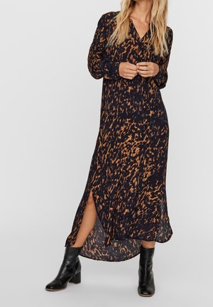 Femme portant une longue robe noire et marron à motifs avec une fente sur le côté et des bottines noires contre un fond uni.