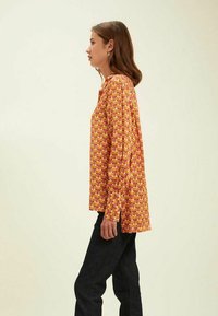 Chemise à manches longues avec un motif floral orange et jaune, en tissu léger, col rond, coupe décontractée, portée avec un jean noir.