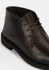 Bottines en cuir marron avec une finition lisse, bout rond et laçage à l'avant. Caractéristiques d'une semelle en caoutchouc noir solide et de coutures minimalistes.