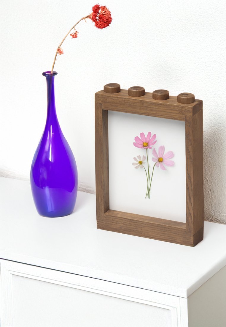 Holzbildrahmen mit abgerundetem oberen Teil, der gepresste rosa Blumen enthält. Neben ihm steht eine hohe, lila Glasvase auf einer weißen Oberfläche.