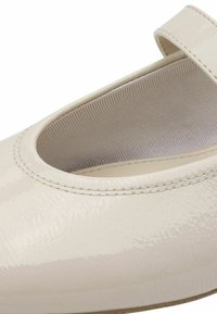 Zapato de charol beige con punta redonda y una sola correa sobre el empeine, que presenta una superficie lisa y brillante con costuras sutiles.