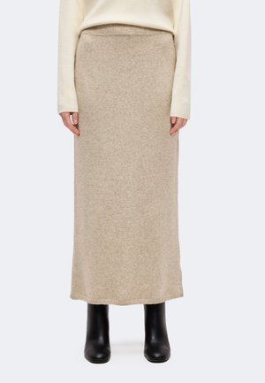Un mannequin porte une longue jupe en maille beige, un pull ivoire et des bottines noires sur un fond clair uni.