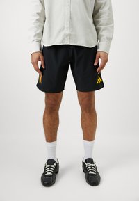 Melni sporta šorti ar dzelteno Adidas logotipu un karoga grafiku kreisajā pusē. Valkāti ar baltām zeķēm un melnām kurpēm.