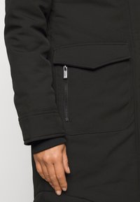 Abrigo acolchado negro con bolsillo con cremallera y bolsillo con solapa. Presenta puños acanalados y una textura suave y mate.