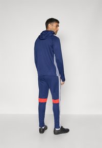 Marineblauwe sportieve hoodie met witte strepen en bijpassende broek met rode accenten; zacht materiaal, slim fit, verstelbare capuchon en sportieve schoenen.