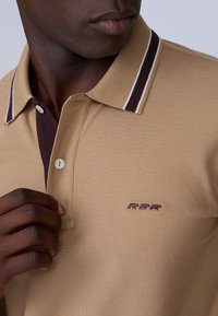 Polo beige en maille texturée, col brun foncé contrastant avec des rayures blanches, et un petit logo éléphant bordeaux sur la poitrine.