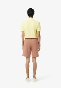 Se lleva una camiseta polo amarilla y unos pantalones cortos terracota, con un ajuste relajado y una textura suave. El look se completa con zapatillas blancas.