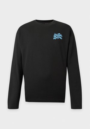 Homme portant une chemise noire à manches longues avec un texte bleu, un jean bleu clair à jambes larges et des chaussures marron, debout devant un fond blanc uni.