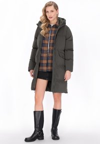 Olívazöld pufi kabát kapucnival, hosszú kivitelben, oldal zsebekkel és cipzáras záródással. Kockás inggel és fekete csizmával viselve.