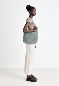Un grand sac tissé de couleur bleu clair, doux et souple, avec une forme arrondie, doté d'une large bandoulière et d'une surface texturée. Un pantalon beige à rayures verticales slim et des sandales noires à plateforme.