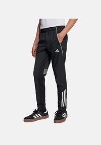 Schwarze Sporthose mit elastischem Bund, weißen Streifen an den Beinen und einem geometrischen Muster an der Seite, getragen mit schwarzen Schuhen.