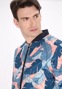 Jachetă bomber cu fermoar frontal, având o bază roz și un model mare de frunze în nuanțe de albastru și alb. Material moale cu guler închis la culoare.