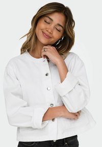 Veste en denim blanc avec un col arrondi, des boutons-pression et des manches retroussées. Elle présente deux poches avant et une coupe légèrement raccourcie.