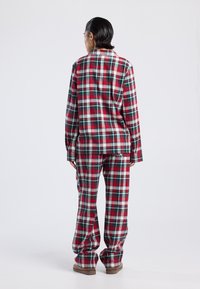 Person standing facing away, wearing red, green, and white plaid pajamas with long sleeves and matching pants on a plain background.