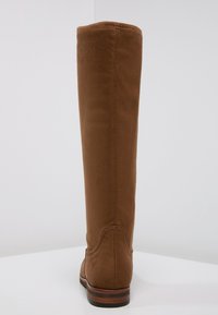 Botte haute en daim marron avec une texture lisse, bout arrondi et talon bas empilé. Caractérisée par des coutures minimales et une finition douce.