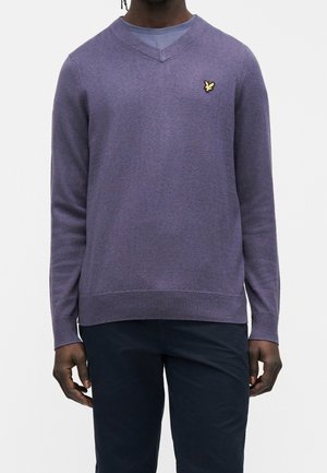 Mann trägt einen violetten V-Ausschnittpullover mit kleinem gelbem Vogel-Logo auf der Brust und dunkelblauen Hosen, steht vor weißem Hintergrund.