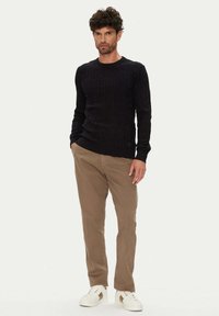 Schwarzer Strickpullover mit Zopfmuster, tailliert geschnitten, mit Rippbündchen an Ärmeln und Saum, kombiniert mit hellbraunen Hosen und weißen Sneakern mit dezentem Logo.