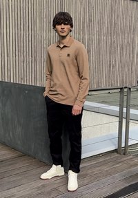 Young man in beige long-sleeve polo, black pants, and white sneakers stands on wooden deck with metal railing and vertical wood panel wall behind.