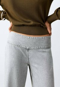 Persona con jeans de tiro alto de color gris claro y un suéter verde oliva, con las manos apoyadas en la parte baja de la espalda, recortada a la altura de la cintura.