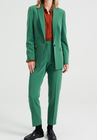 Grön blazer och byxor i en slät textur. Brun skjorta under. Svarta loafers på fötterna. Rent snitt och skräddarsydd passform.