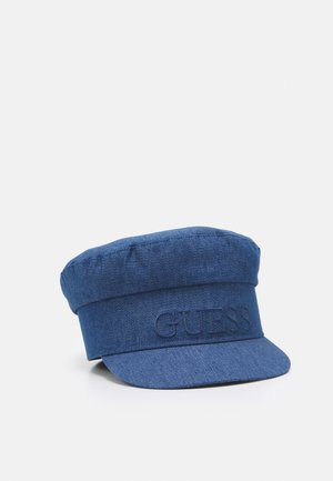 Blauwe denim pet met vlakke bovenkant, korte klep en verhoogd "GUESS" logo op de voorste band, getoond tegen een witte achtergrond.