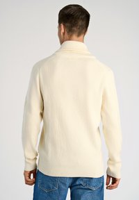 Cremfarbener gerippter Strickpullover mit hohem Kragen und langen Ärmeln. Der Stoff hat ein strukturiertes Muster und wird mit blauen Jeans kombiniert.