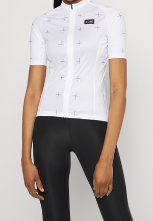 Kobieta ubrana w dopasowaną białą koszulkę kolarską z małymi czarnymi krzyżykami i czarne obcisłe legginsy, stojąca z rękami swobodnie opuszczonymi wzdłuż boków.