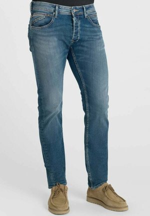Man met een slim-fit blauwe spijkerbroek en lichtbruine casual suède schoenen, staand tegen een effen achtergrond.