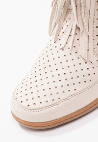 Chaussure en daim beige avec perforations carrées, lacets à franges et semelle en caoutchouc marron clair, présentée en gros plan sur fond blanc.