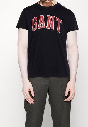 Person trägt ein schwarzes T-Shirt mit kurzen Ärmeln mit großem roten "GANT"-Schriftzug und dunkelgraue Hosen, steht vor einem schlichten weißen Hintergrund.