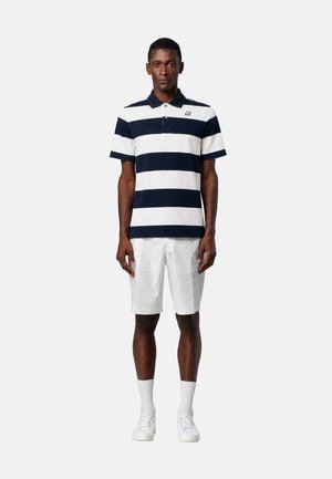 Uomo in piedi che indossa una polo a righe orizzontali blu navy e bianche, pantaloncini bianchi, calzini bianchi e scarpe da ginnastica bianche su uno sfondo semplice.