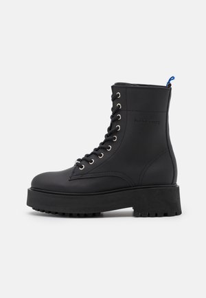 Bota negra de cuero con cordones, suela de plataforma gruesa, ojales plateados y una lengüeta azul en la parte trasera. Textura lisa y diseño estructurado.