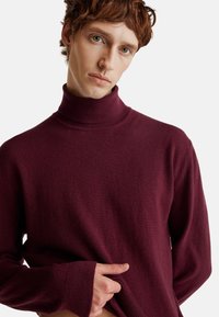 Bordowy sweter z golfem wykonany z miękkiej tkaniny. Posiada wysoki kołnierz, ściągacze przy rękawach oraz luźny krój dla komfortu.