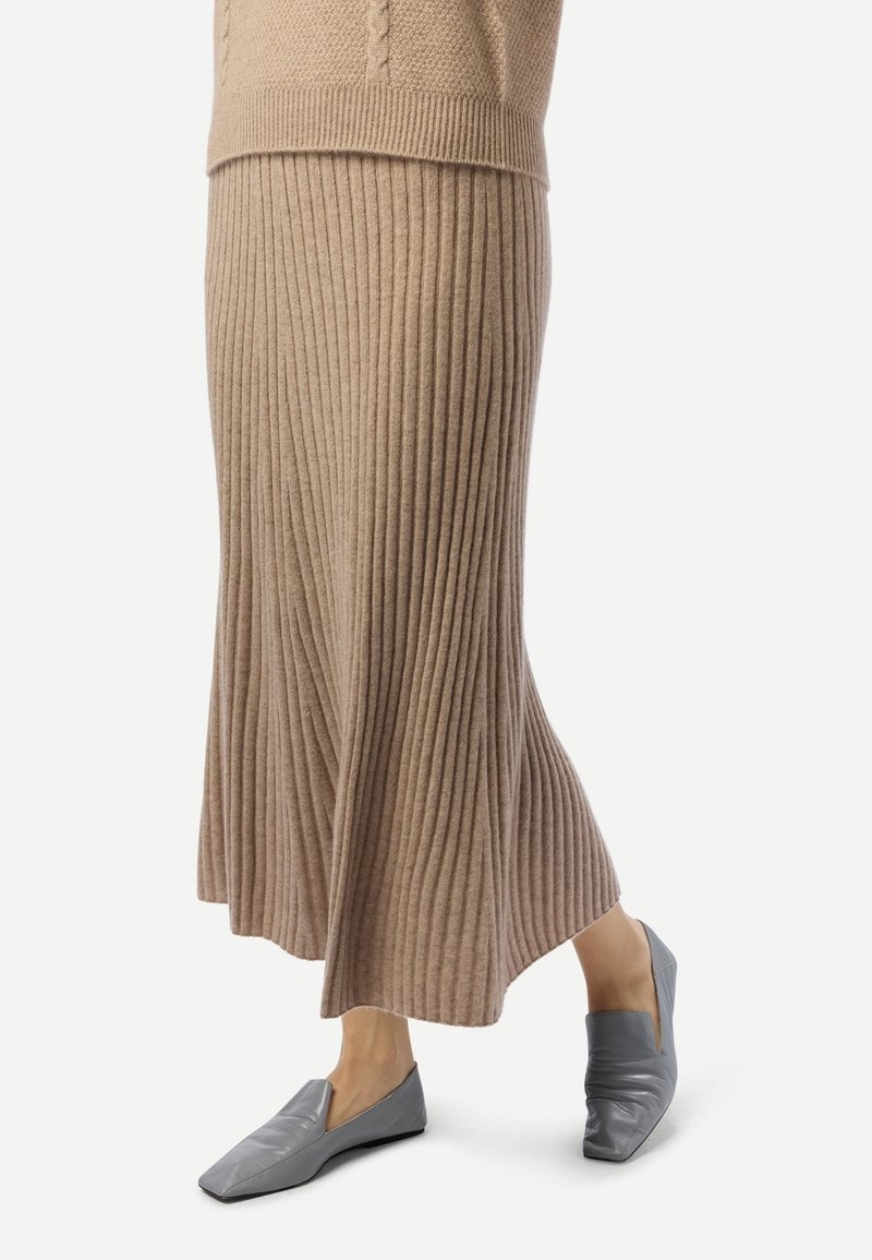 Jupe midi en maille côtelée beige associée à des chaussures plates grises à enfiler, portée par une personne debout sur un fond blanc.