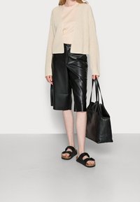 Beige gestrickte Strickjacke, beiges Hemd, schwarze Shorts aus Kunstleder, schwarze Sandalen und eine schwarze Ledertasche. Glatte Texturen und lässiger Stil.