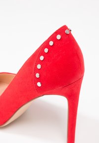 Escarpin rouge en suede avec un bout pointu et des clous en argent décoratifs le long du talon arrière, doté d'une semelle intérieure lisse beige.