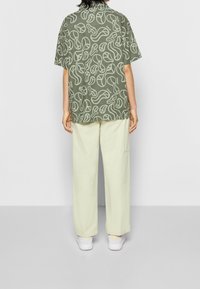 Camicia verde a maniche corte con motivi astratti, abbinata a pantaloni a gamba larga color beige chiaro. L'outfit è completato da sneakers bianche.