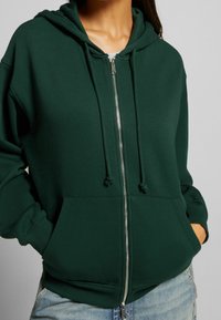 Felpa verde con zip realizzata in tessuto morbido, dotata di cappuccio regolabile con cordini, tasche frontali e polsini a coste. Design semplice con una vestibilità comoda.