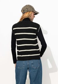 Pull sans manches noir et blanc rayé, texture côtelée, porté par-dessus un haut à manches longues noir, associé à un jean taille haute bleu clair.