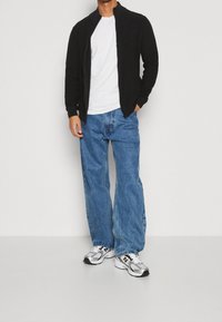 Veste noire à fermeture éclair, t-shirt blanc, jean bleu à jambes larges, et baskets grises avec des éléments blancs. Design de tenue simple et décontractée.