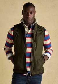 Olivgrüne gesteppte Weste über einem mehrfarbigen gestreiften Hemd mit roten und blauen Streifen. Verfügt über Reißverschlusstaschen und einen Stehkragen.