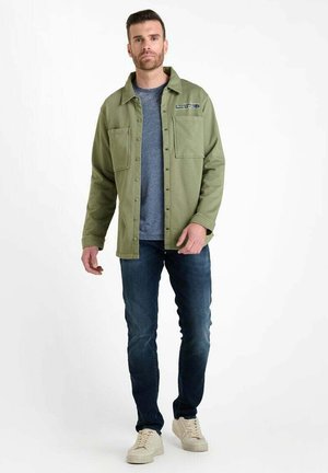 Mann trägt ein olivgrünes Hemd über einem blauen T-Shirt, dunkle Jeans und beige Sneaker und steht vor einem weißen Hintergrund.