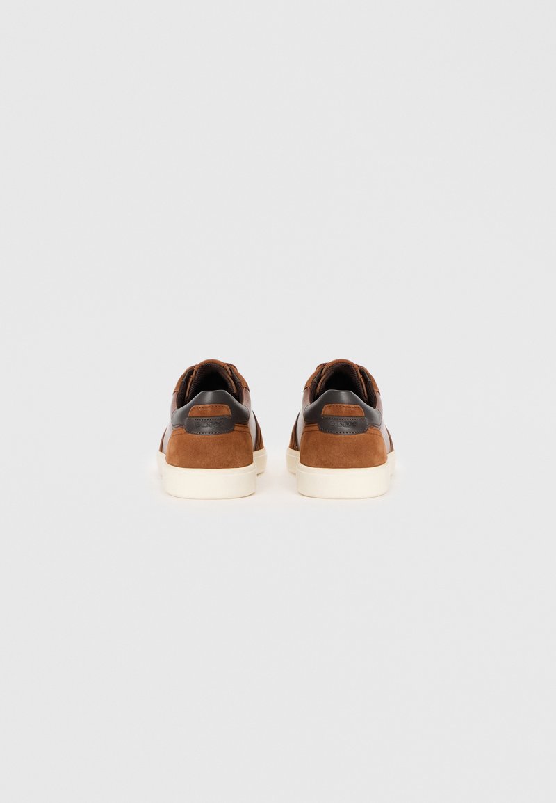 Baskets en daim et cuir marron avec une semelle plate couleur crème, présentant un design cousu et un accent de logo sur le talon.