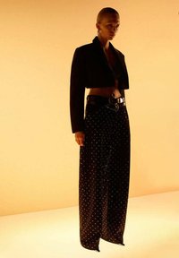 Mujer vestida con una chaqueta negra corta, pantalones de pierna ancha de cintura alta con pequeños puntos blancos, un cinturón ancho y grandes pendientes contra un fondo beige.