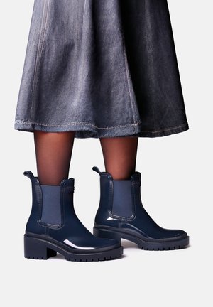 Personne portant des collants noirs transparents, une jupe en jean longueur genou et des bottines bleu marine brillantes à talons blocs.
