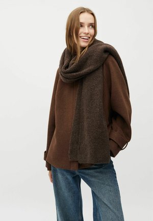 Brun oversized sweater med brede ærmer, parret med et mørkebrunt ribbet tørklæde. Modellen har afslappede blå jeans på. Glat tekstur hele vejen igennem.
